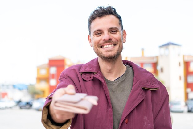 Young caucasian man at outdoors holding a wallet with happy expression