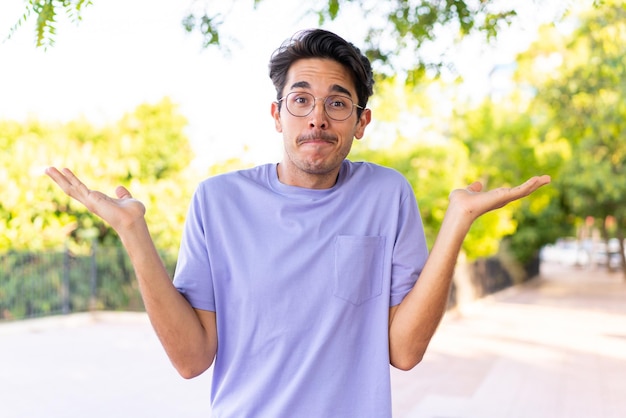 Young caucasian man at outdoors in a park having doubts while raising hands