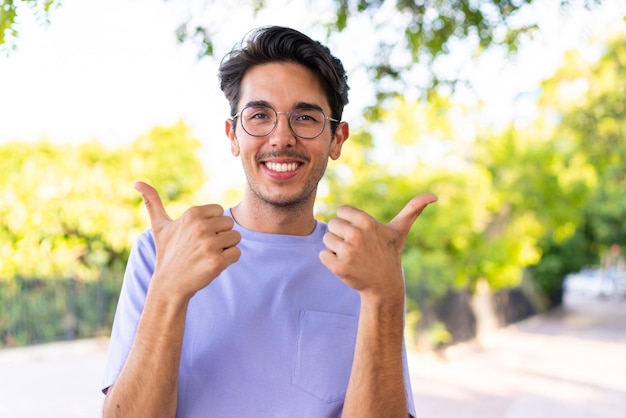 Young caucasian man at outdoors in a park with thumbs up gesture and smiling