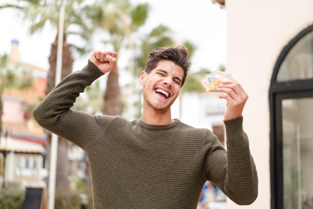 Photo young caucasian man at outdoors taking a lot of money and celebrating a victory