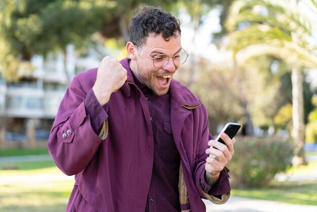 Young caucasian man at outdoors using mobile phone and doing victory gesture