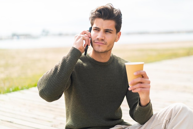 Young caucasian man at outdoors using mobile phone and holding a coffee