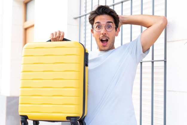 Young caucasian man at outdoors in vacation with travel suitcase and surprised