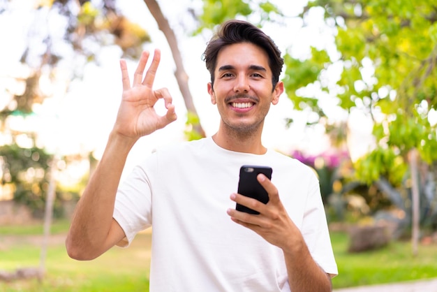 Young caucasian man in a park using mobile phone and doing OK sign