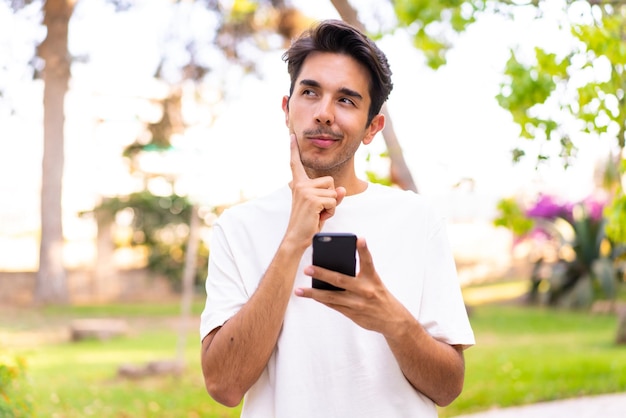 Young caucasian man in a park using mobile phone and thinking