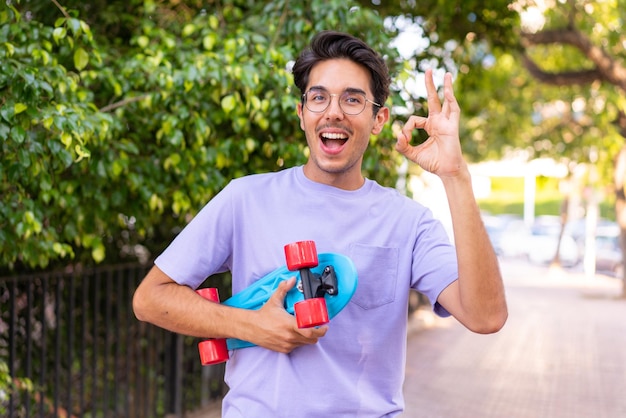 Young caucasian man in a park with a skate and doing OK sign