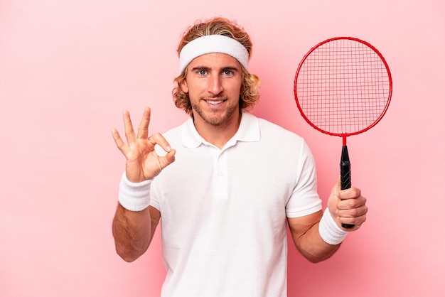 Young caucasian man playing badminton isolated on pink background cheerful and confident showing ok gesture.