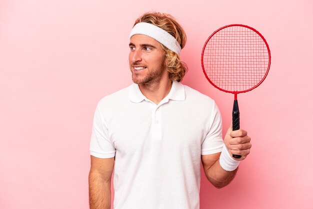 Young caucasian man playing badminton isolated on pink background looks aside smiling, cheerful and pleasant.