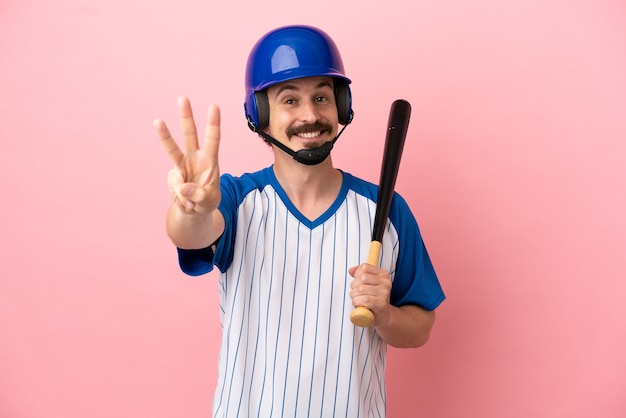 Young caucasian man playing baseball isolated on pink background happy and counting three with fingers