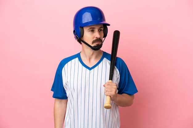 Young caucasian man playing baseball isolated on pink background looking to the side