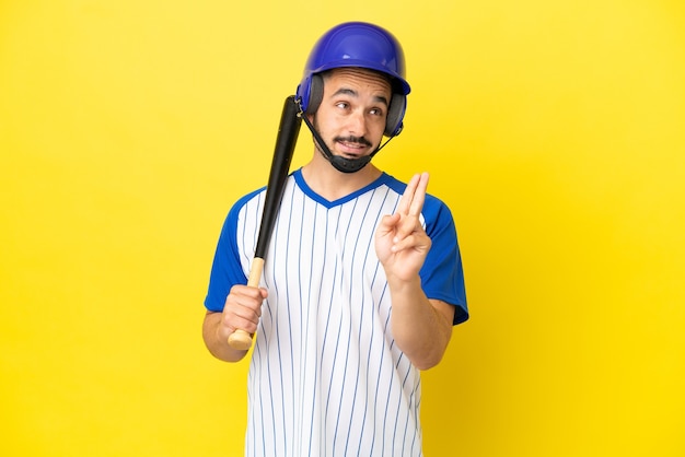 Young caucasian man playing baseball isolated on yellow background with fingers crossing and wishing the best