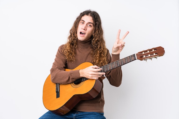 Young caucasian man playing guitar isolated joyful and carefree showing a peace symbol with fingers.