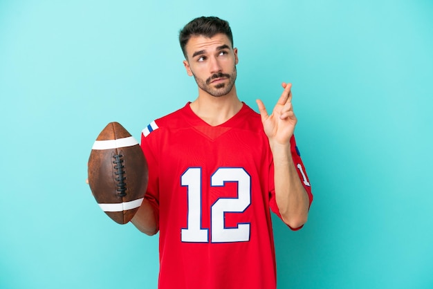 Young caucasian man playing rugby isolated on blue background with fingers crossing and wishing the best