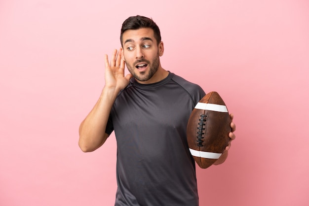 Young caucasian man playing rugby isolated on pink background\
listening to something by putting hand on the ear