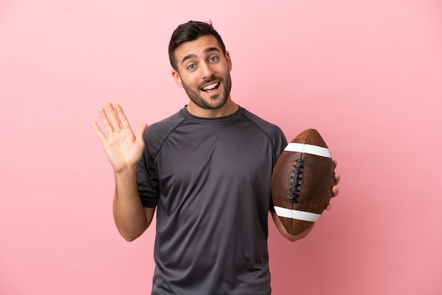 Young caucasian man playing rugby isolated on pink background saluting with hand with happy expression