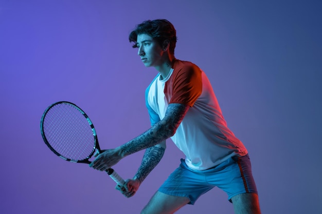 Молодой кавказец, играющий в теннис, изолированный на пурпурно-голубом студийном фоне в неоновом действии и концепции движения