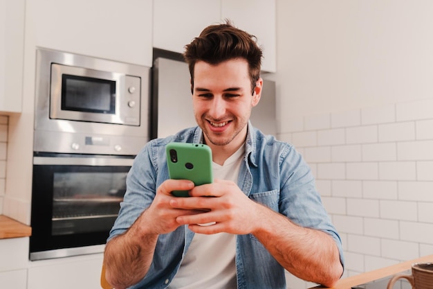 Young caucasian man playing a video game with a cellphone sitting at home kitchen Happy guy using a smartphone app to send messages on social media Teenage male smiling browsing with a mobile phone