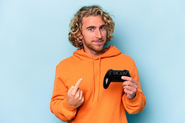 Фото Молодой кавказский мужчина играет с контроллером видеоигры, изолированным на синем фоне, указывая пальцем на вас, как будто приглашая подойти ближе.