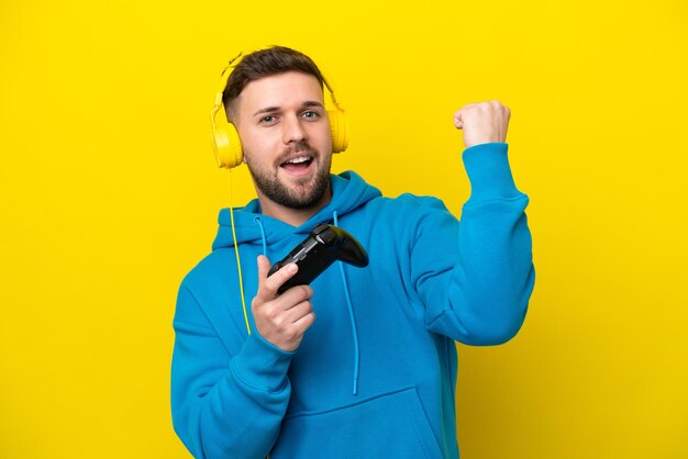 Young caucasian man playing with a video game controller isolated on yellow background celebrating a victory