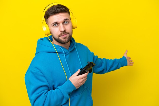 Young caucasian man playing with a video game controller\
isolated on yellow background extending hands to the side for\
inviting to come
