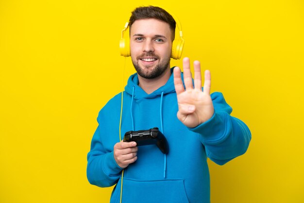 Молодой кавказский мужчина играет с контроллером видеоигры, изолированным на желтом фоне, счастливым и считая четыре пальцами