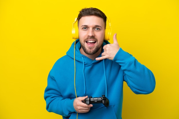 Young caucasian man playing with a video game controller\
isolated on yellow background making phone gesture call me back\
sign