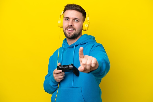 Young caucasian man playing with a video game controller isolated on yellow background showing and lifting a finger