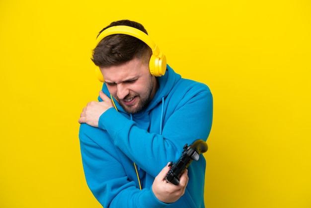 努力をしたために肩の痛みに苦しんでいる黄色の背景に分離されたビデオ ゲーム コントローラーで遊ぶ若い白人男性