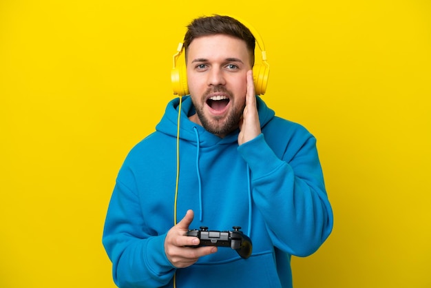 Young caucasian man playing with a video game controller isolated on yellow background with surprise and shocked facial expression