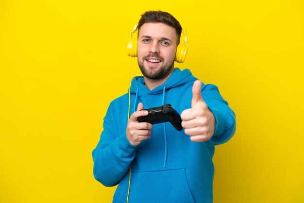 Young caucasian man playing with a video game controller isolated on yellow background with thumbs up because something good has happened