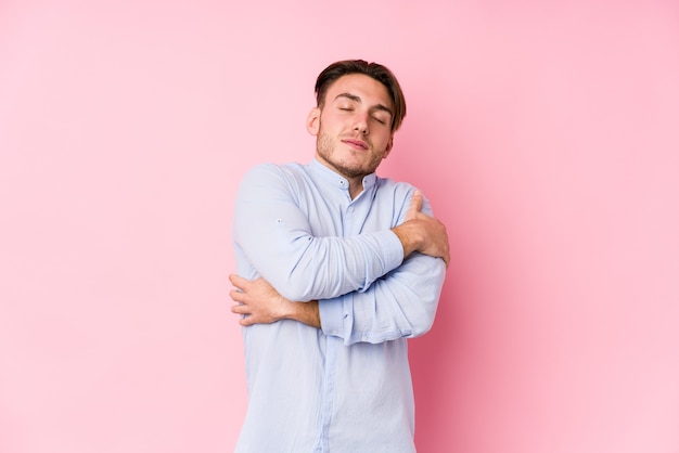 Giovane uomo caucasico in posa in un muro rosa isolato abbracci, sorridente spensierato e felice.