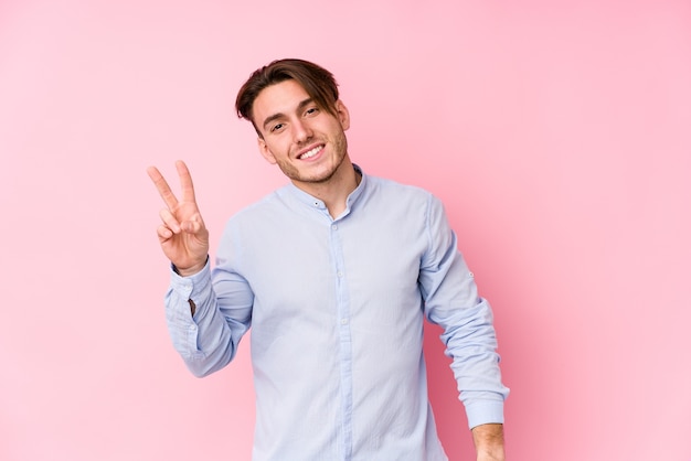 Young caucasian man posing in a pink wall isolated joyful and carefree showing a peace symbol with fingers.