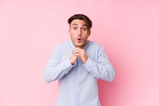 Young caucasian man posing in a pink wall praying for luck, amazed and open mouth