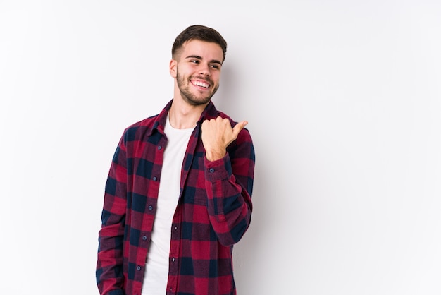 Young caucasian man posing in a white wall isolated points with thumb finger away, laughing and carefree.