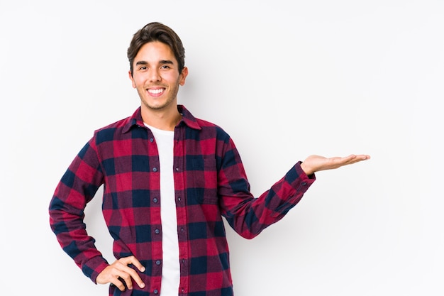 Young caucasian man posing in a white wall showing a blank space on a palm and holding another hand on waist.