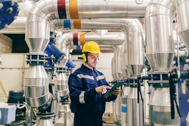 Young Caucasian man in protective suit using tablet while standing in heating plant.