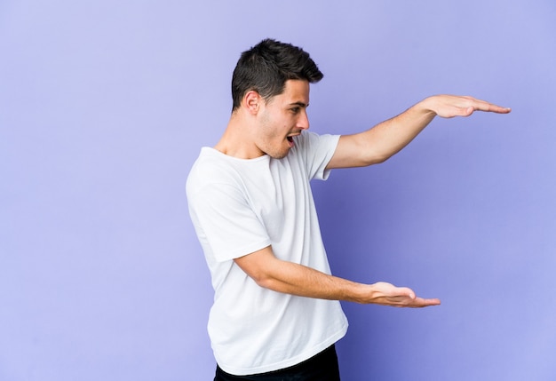 Young caucasian man on purple shocked and amazed holding a copy space between hands.