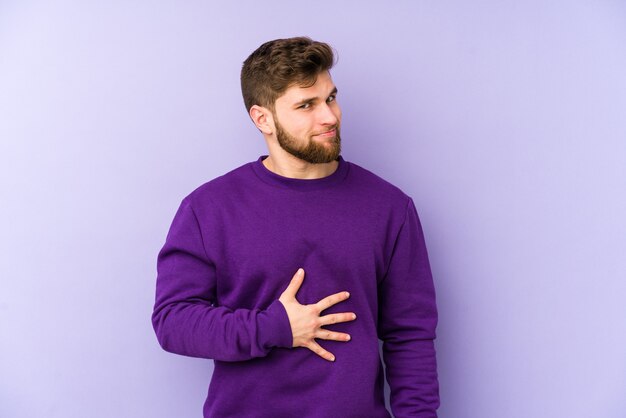 Young caucasian man on purple touches tummy, smiles gently, eating and satisfaction concept.