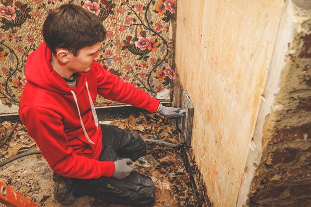 Photo young caucasian man in a red hoodie squatting removes retro wallpaper on a steamboat in an old house