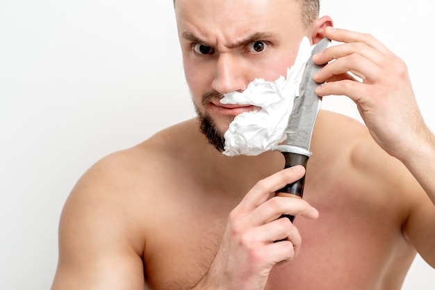 Foto giovane uomo caucasico che rade la barba con il coltello su sfondo bianco