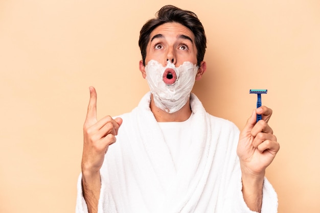 Young caucasian man shaving his beard isolated on beige background pointing upside with opened mouth.