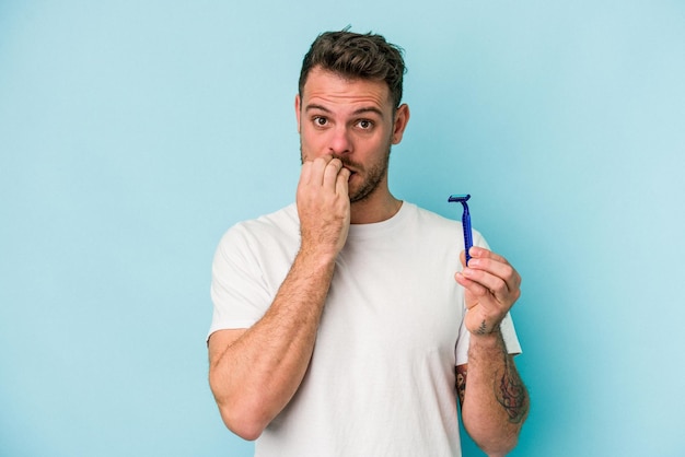 Young caucasian man shaving his beard isolated on blue background biting fingernails nervous and very anxious