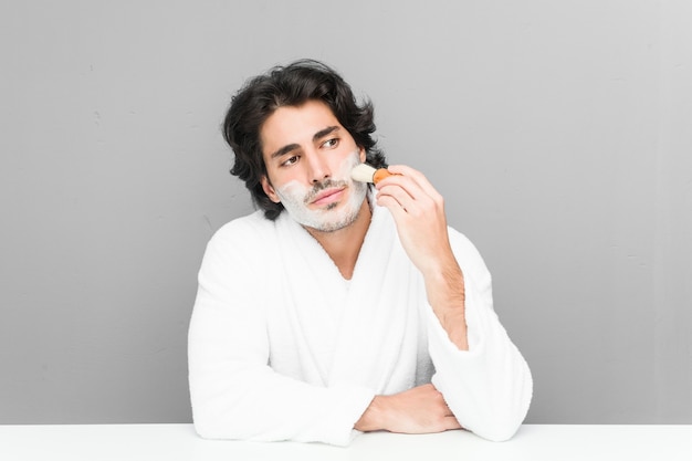 Young caucasian man shaving his beard isolated on a grey background