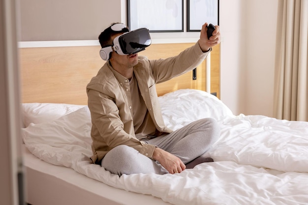 Giovane uomo caucasico seduto sul letto a casa con auricolare vr e giocare a videogiochi interattivi esplorando la realtà virtuale uomo che indossa occhiali vr gadget futuri tecnologia educazione studio online