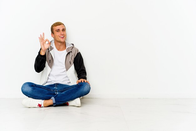 Young caucasian man sitting on the floor cheerful and confident showing ok gesture.