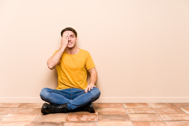 Young caucasian man sitting on the floor having fun covering half of face with palm.