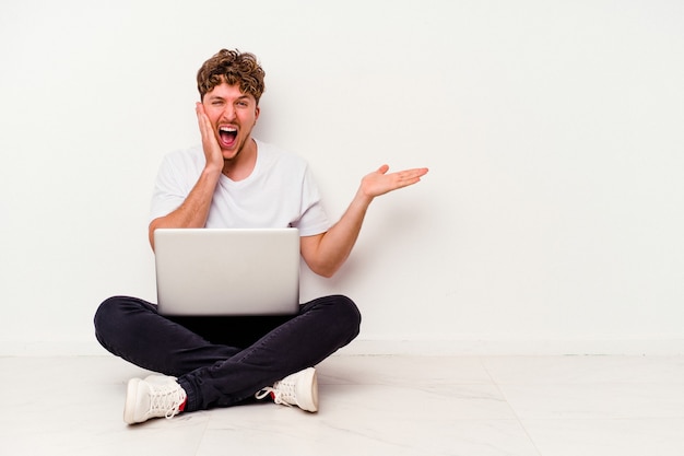 Young caucasian man sitting on the floor holding on laptop isolated on white background holds copy space on a palm, keep hand over cheek. Amazed and delighted.