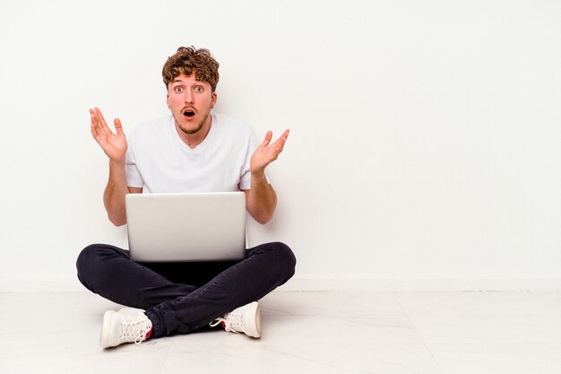 Young caucasian man sitting on the floor holding on laptop isolated on white background surprised and shocked.