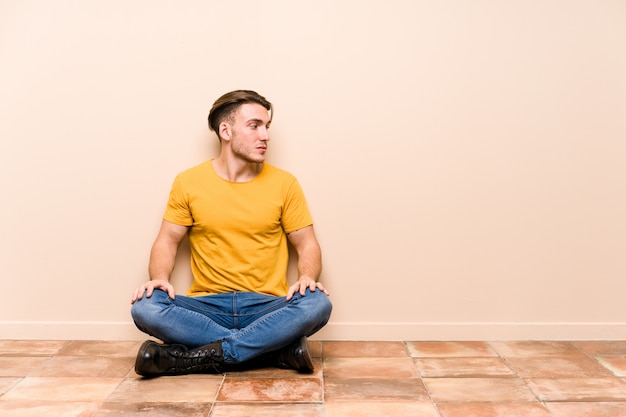 Young caucasian man sitting on the floor isolated gazing left, sideways pose