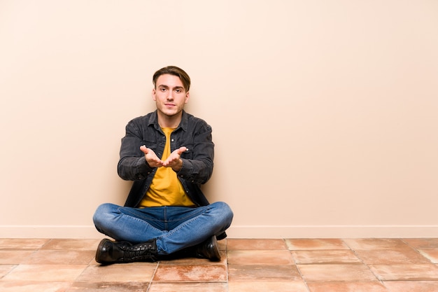 Foto il giovane uomo caucasico che si siede sul pavimento ha isolato la tenuta qualcosa con la palma